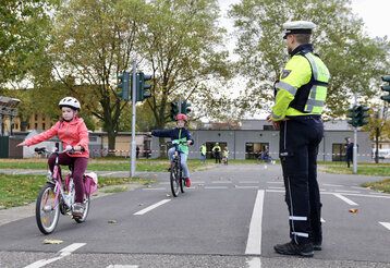 zwei Kinder fahren auf dem Verkehrsübugsplatz mit den Fahrrädern, Polizist guckt zu