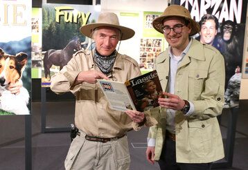 Zwei Männer stehen  in Safari-Anzügen im Filmmuseum vor alten Filmplakaten, in der Hand ein Buch über Lassie