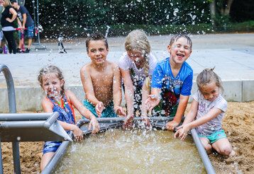 5 Kinder spielen an einer Wasseranlage auf einem Spielplatz, Wasser spritzt, die Kinder lachen