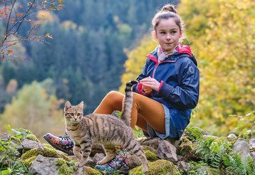 Ein Mädchen sitzt in der Natur auf einem Stein, im Hintergrund Wald und im Vordergrund eine Katze