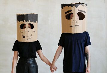 Eine Frau und ein Mann stehen nebeneinander und halten sich an den Händen, beide tragen Karton-Masken mit Frankenstein-Gesichtern bemalt