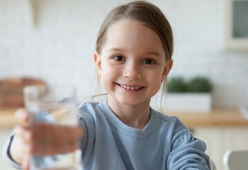 kleines Mädchen lächelt und hält ein Wasserglas in die Kamera